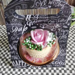 A clear glass dome containing a pink rose soap. The dome is placed on a black and white chequered surface. The dome is surrounded by a black box with white lettering that reads "Love Wash Dry", "Every Moment Only Soap", "Enjoy the Little Things", and "Today is a Good Day".