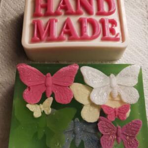 A green soap bar with a white bar on top. The white bar has the word "HANDMADE" embossed in pink letters. The green bar has several pink, white, and blue butterfly decorations.