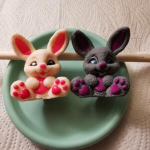 Two bunny figurines, one cream-coloured and one grey, sit on a light green plate. The bunnies have pink inner ears, noses, and paw prints. A wooden dowel rests behind them. The figurines are likely decorative items or soap.