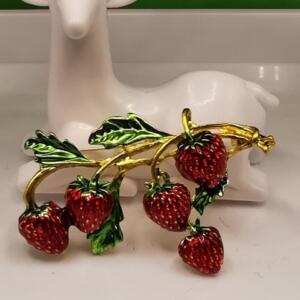 A gold-tone brooch featuring a cluster of red enamel strawberries on a green enamel vine. The strawberries have a shiny finish and are arranged in a cascading design.