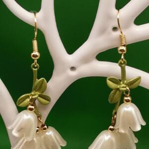 A pair of gold dangle earrings with white flower charms and green leaves. The flowers are made of a pearlescent material.