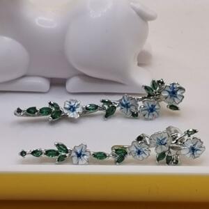 A pair of silver earrings with a floral design. The earrings feature white flowers with blue centres and green leaves.
