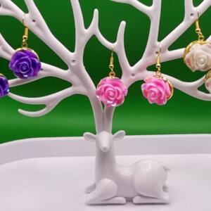 Three pairs of rose-shaped earrings displayed on a white deer-shaped stand. The earrings are in shades of purple, pink, and white.
