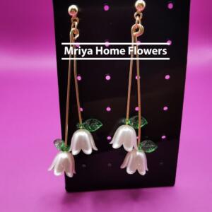 A pair of gold dangle earrings with white flower charms and green leaves. The earrings are displayed on a black card with the text "Mriya Home Flowers".