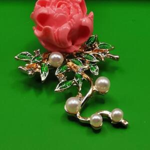 A pink peony flower brooch with gold-tone metal detailing. The brooch has green enamel leaves, faux pearls, and small clear crystals. The design includes a branch with additional pearls.