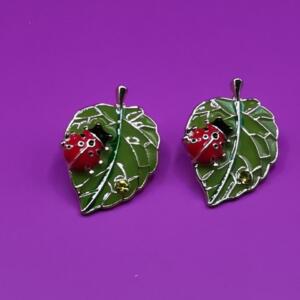 A pair of silver earrings featuring a green leaf design with a red ladybug on each leaf.