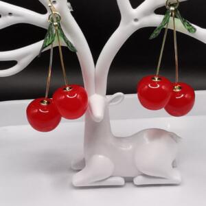 A pair of cherry earrings with green leaves. The earrings are made of red and gold coloured resin and are hanging from a white deer-shaped earring stand.