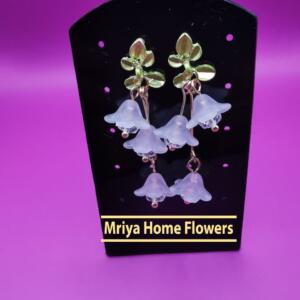 A pair of white flower dangle earrings with gold accents and green leaves. The earrings are displayed on a black stand with the text "Mriya Home Flowers".