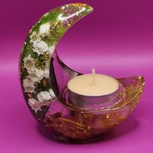 A clear glass crescent moon candle holder with gold and green glitter embedded inside. The holder has a small tea light candle inside.