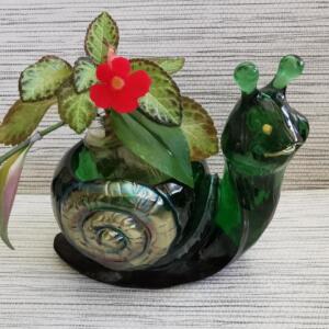 A decorative, dark green glass snail-shaped planter. It contains a small plant with green and brown leaves and a bright red flower. The snail has a spiral shell design and two green antennae. A clear glass jar holds the plant.
