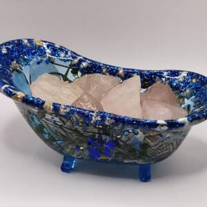 A blue and white resin bathtub with glitter and floral accents, filled with pink rose quartz crystals. The bathtub has three blue feet.