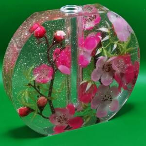 A clear glass vase with a round shape and a glitter finish. The vase is filled with pink flowers and green leaves.