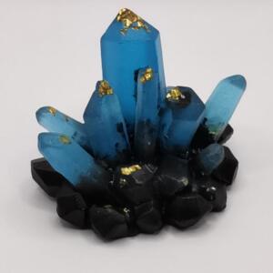 A decorative crystal cluster. The crystals are a bright blue, adorned with gold leaf. The base is made of black crystals. The arrangement is natural and organic. The crystals are a striking decorative piece.