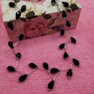 A silver chain necklace with black teardrop-shaped beads. The necklace is on a pink surface.