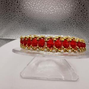 A red and gold beaded bracelet with white pearls. The bracelet is on a clear display stand.