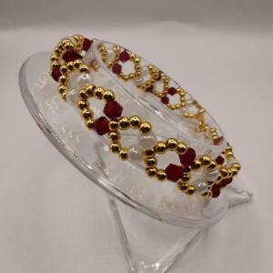 A bracelet featuring gold-coloured, red, and white beads. The bracelet is presented on a clear, angled stand. The beads are arranged in a repeating pattern, creating a decorative accessory.